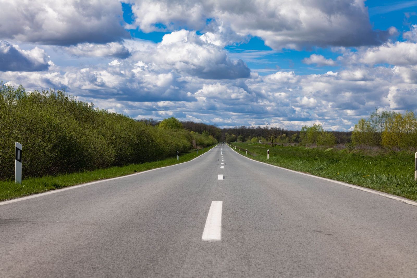 A straight two-lane road between green fields and trees under a cloudy blue sky